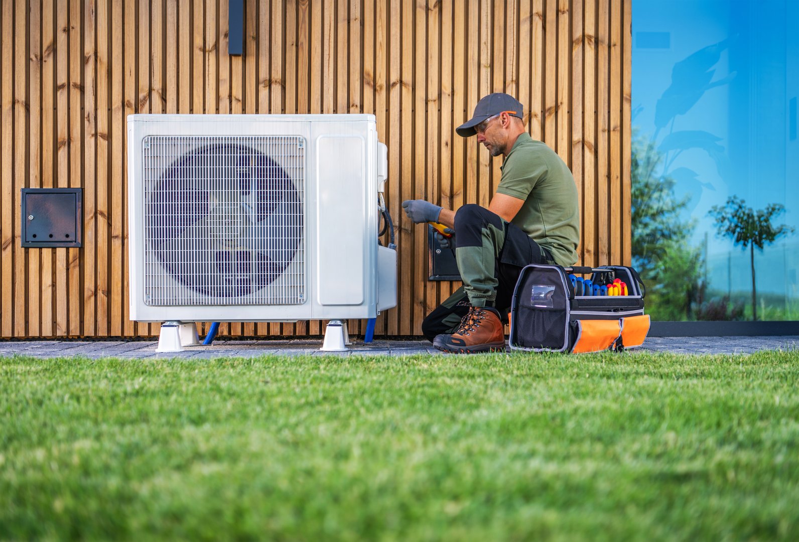 A technician is servicing an air conditioning unit located beside a modern home. Dressed in work attire, he uses tools from an organized toolbox while seated on the grass. The setting features a wooden wall and bright blue windows, indicating a well-maintained property.